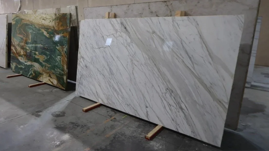 Large marble slabs standing upright on wooden supports in a warehouse, with one slab featuring white and beige streaks and the other slab showing green and gold patterns.