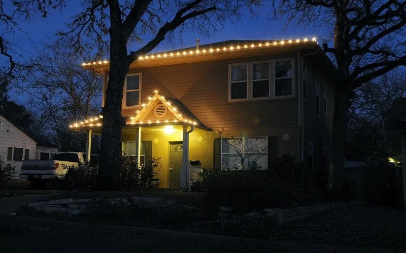 A two-story house decorated with string lights along the roofline and around the front porch at dusk, with trees and neighboring houses visible in the background.