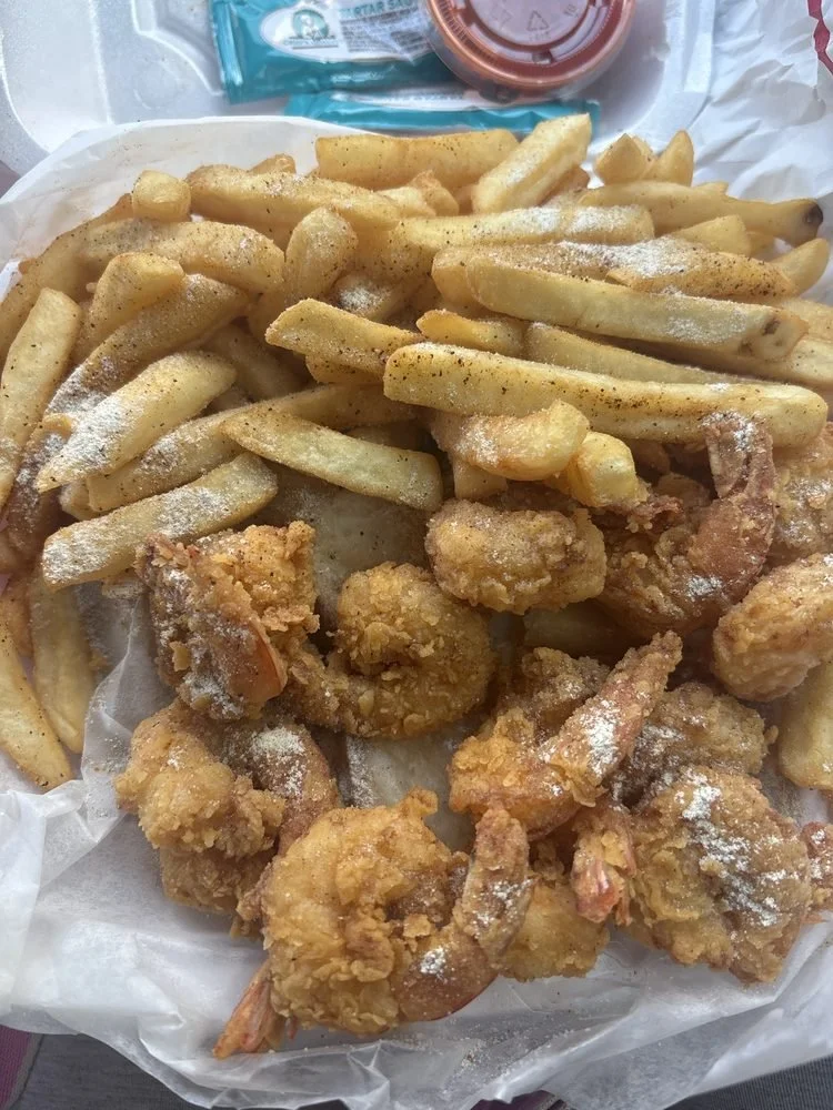 French fries and fried shrimp in a takeout container with condiments in cups in the background.