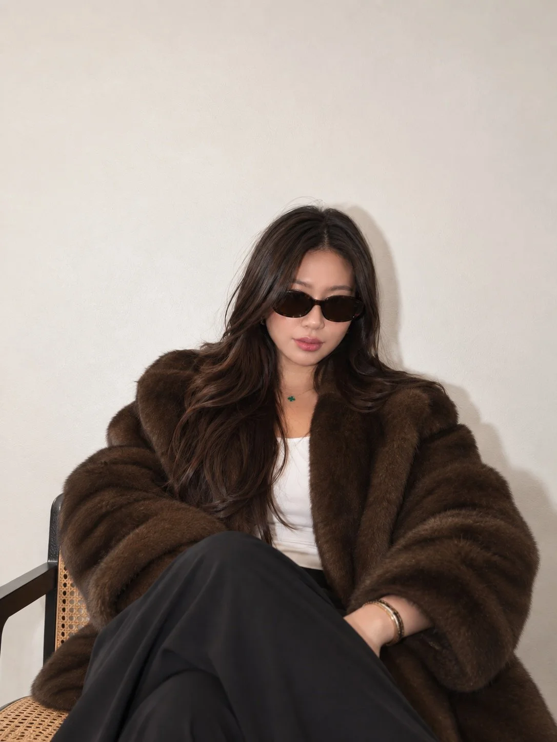 Chic woman in a brown faux fur coat and black sunglasses wearing a minimalist neutral outfit, seated against a cream wall.