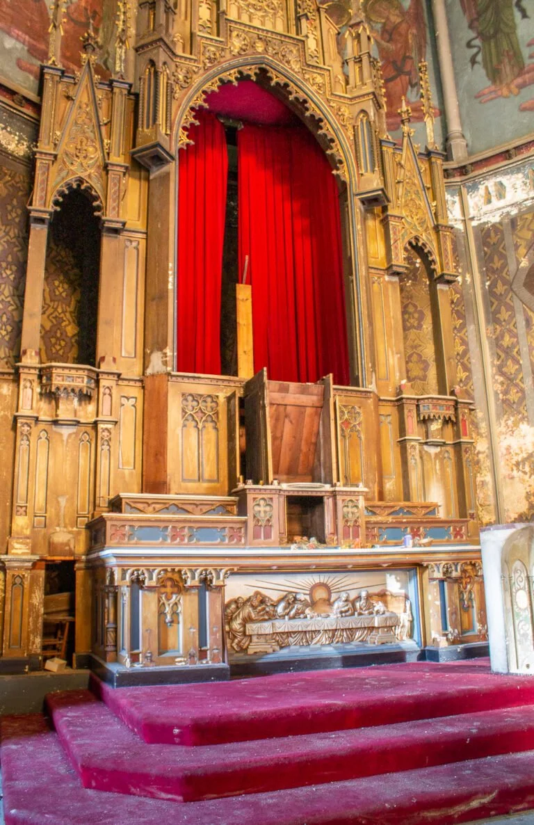 An ornate, wooden church altar with gilded details, red curtains, and a religious relief sculpture of The Last Supper. Red-carpeted steps lead up to the altar in a church interior.