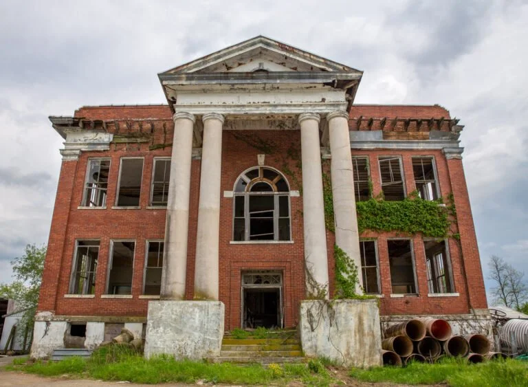 An abandoned two-story brick mansion with broken windows and overgrown vegetation, featuring a large porch with four tall columns and a triangular pediment.