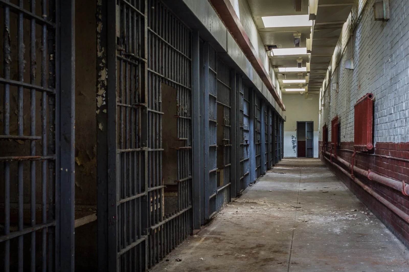 Empty jail corridor with metal cell doors on the left and red brick wall with pipes on the right.