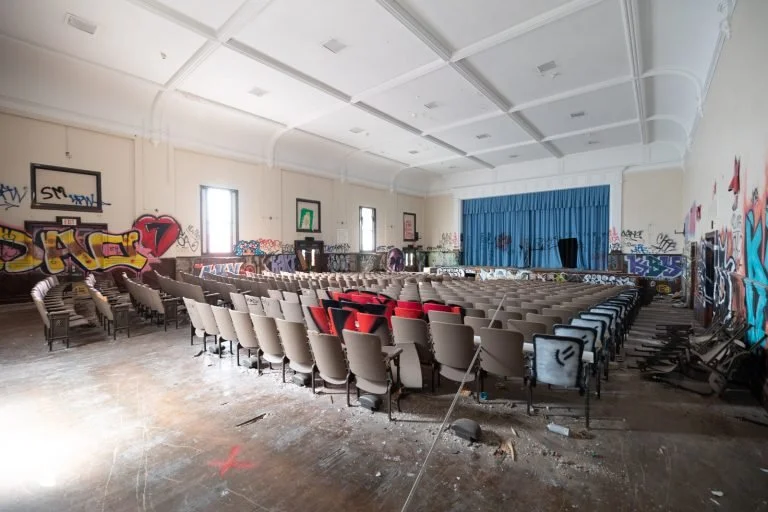 An abandoned auditorium with graffitied walls, a stage with a blue curtain, and rows of beige chairs, some stacked or knocked over.
