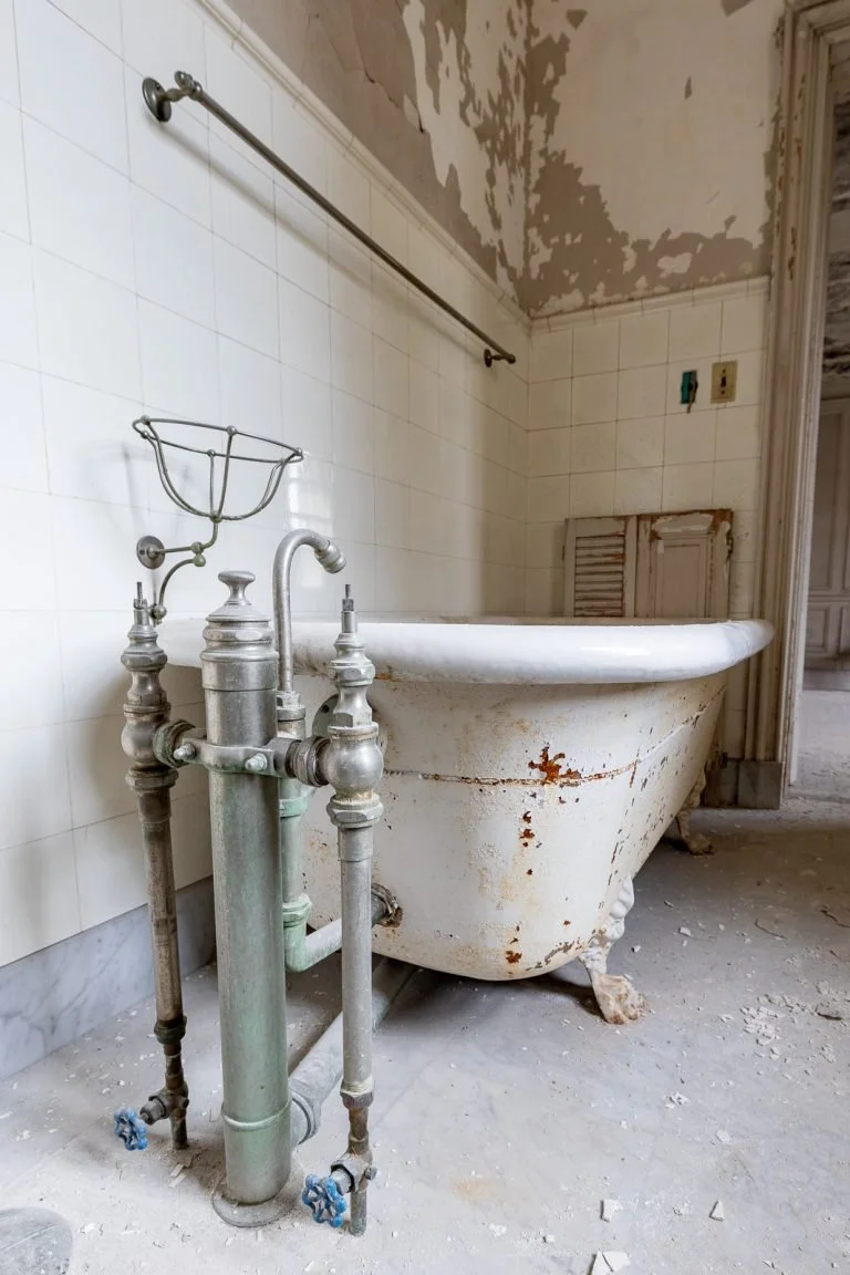 Old, rusty clawfoot bathtub with peeling paint in a dilapidated bathroom with peeling ceiling paint and dirty floor.