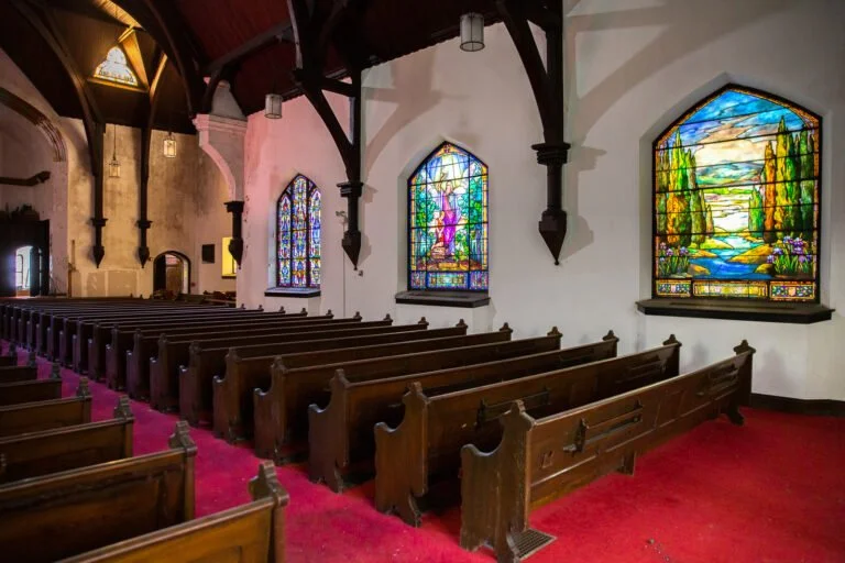 Interior of a church with wooden pews, stained glass windows depicting religious scenes, and a red carpeted floor.