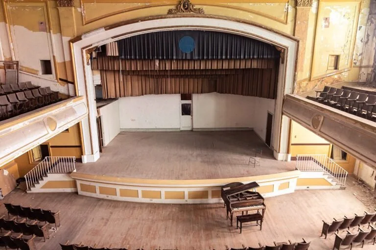 Empty stage with a black piano, in an old theater with yellow walls and dark wood floors, viewed from the audience area.