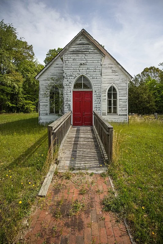 An old, weathered white church with a red door and arched windows, accessed by a wooden ramp on a grassy field.