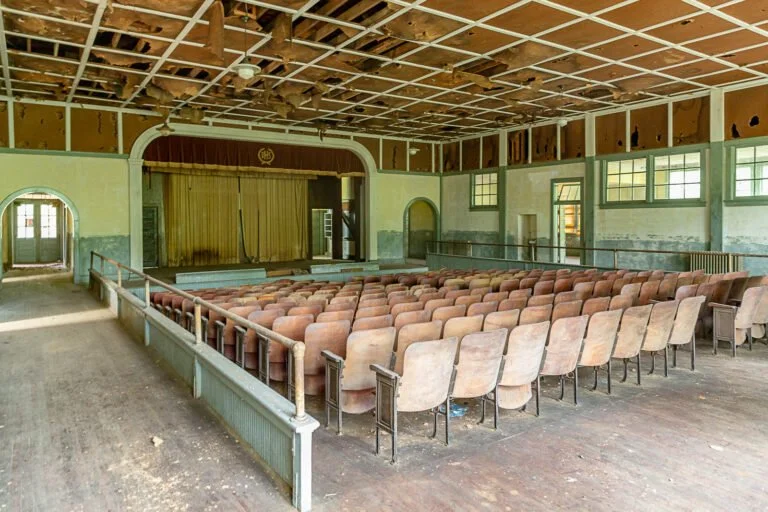 Empty auditorium with a stage and rows of old, worn chairs, peeling walls, and a damaged ceiling.