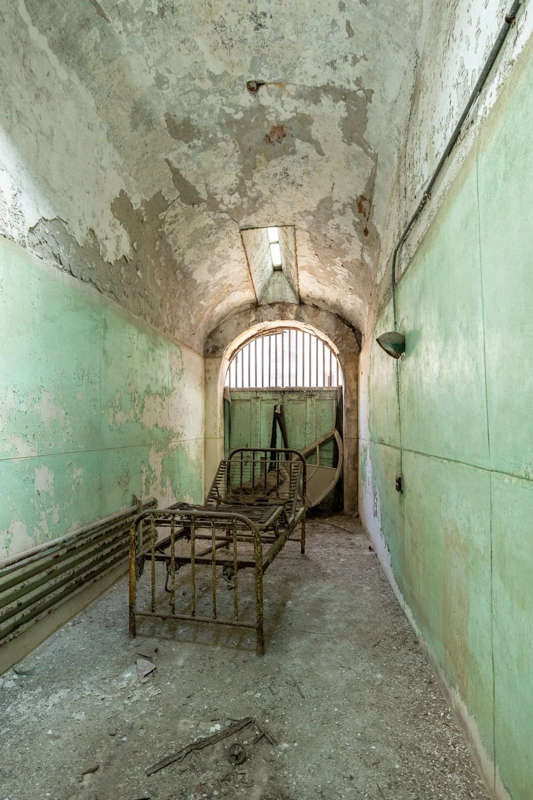 An abandoned hospital corridor with peeling paint, a rusty hospital bed, and broken equipment inside a deteriorating building.