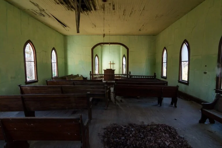 Interior of an abandoned church with green walls, wooden pews, stained glass windows, and a small altar at the front. The ceiling is damaged and peeling.