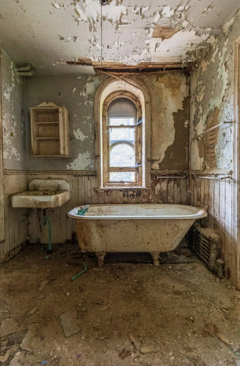 Abandoned bathroom with a rusted bathtub, a sink, peeling paint, broken ceiling, dirty floor, and a small window.