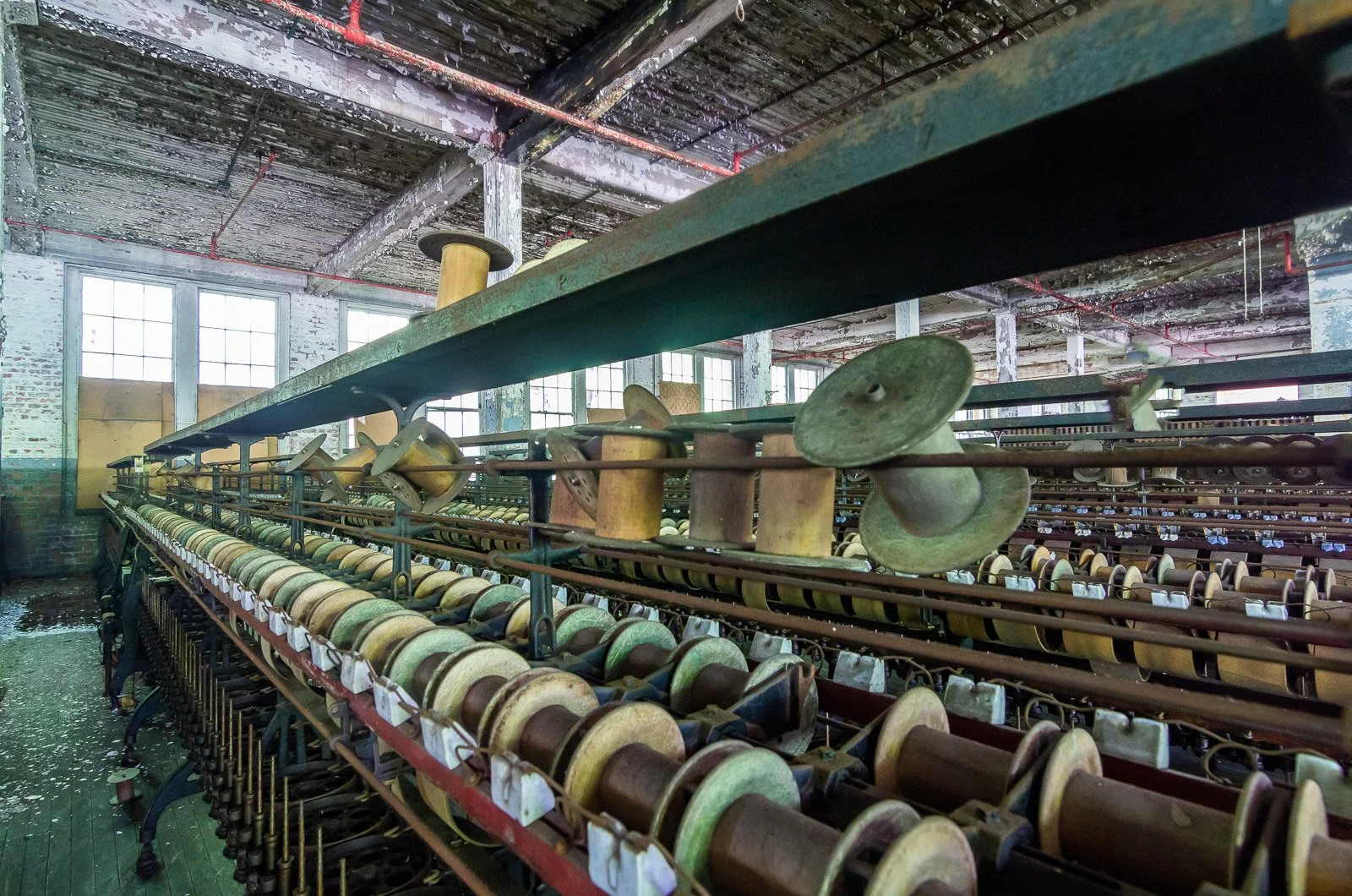 Industrial workshop with rows of old bobbins and spools on metal racks in a rustic, abandoned factory with large windows and peeling brick walls.