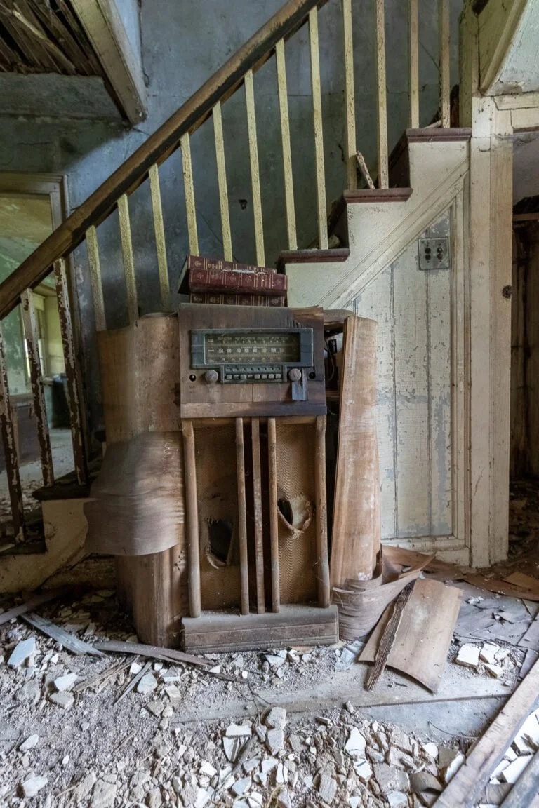 Old, dusty wooden radio and bookshelf in abandoned, debris-filled house with peeling paint and damaged stairs.