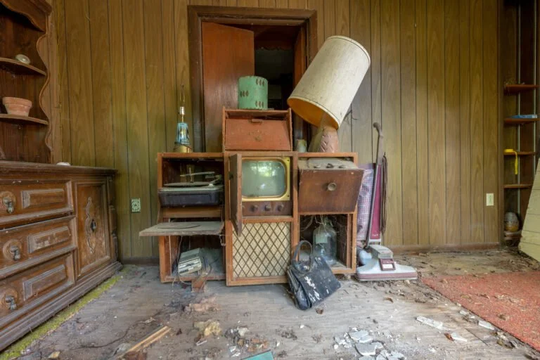 A cluttered antique room with a wooden wall, old furniture, a vintage television, a tilted lampshade, and debris on the floor.