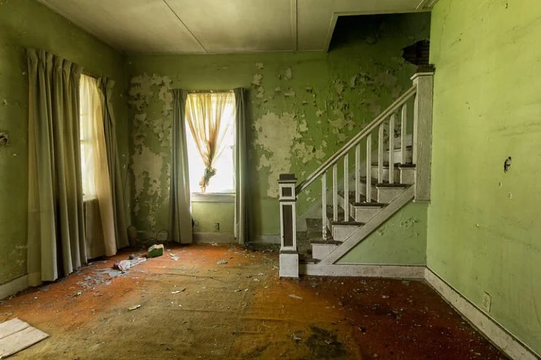 An abandoned room with green peeling walls, a worn carpet, multiple damaged curtains, and a staircase leading upstairs.