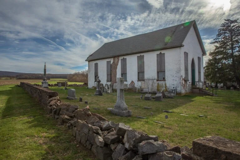 An old, weathered white church with boarded-up windows and a pitched roof, surrounded by a graveyard with broken and upright headstones, a stone wall in the foreground, and a cloudy sky overhead.