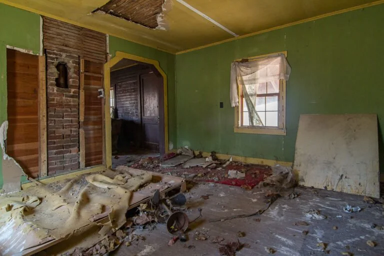 An interior room with peeling paint, damaged ceiling, and debris scattered on the floor, including insulation, wood, and trash. The room has a window with broken or missing curtains and an open doorway leading to another dark room.