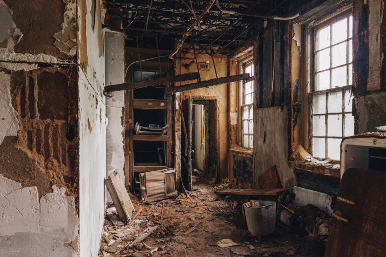 Interior of a dilapidated, abandoned house with damaged walls, broken windows, and debris on the floor.