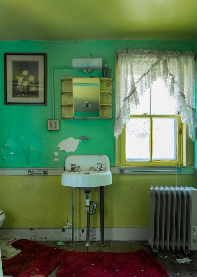 An old, worn bathroom with peeling green and yellow paint, a small wall-mounted sink with two faucets, a radiator, a window with lace curtains, and a vintage mirror cabinet. The room appears neglected and in disrepair.