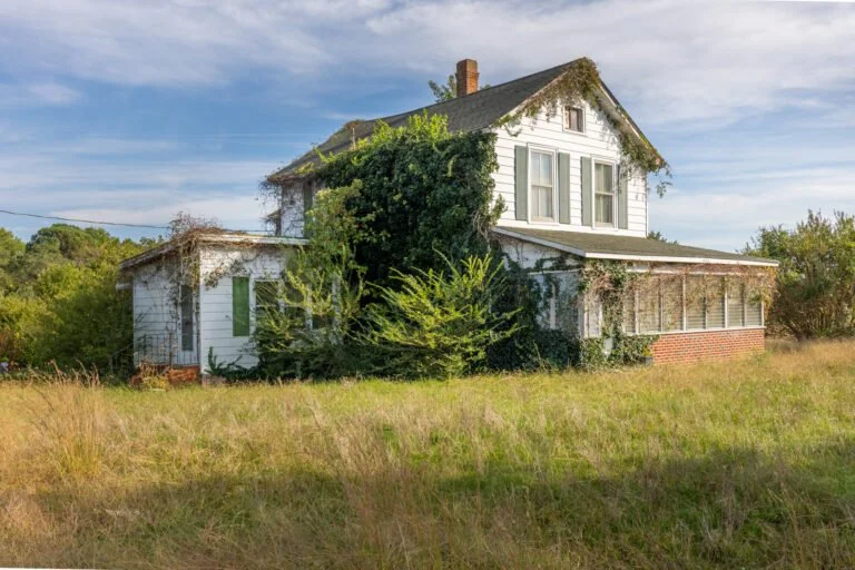 An old, abandoned house with overgrown bushes and grass in front. The house has a white exterior, a sloped roof with moss, and multiple windows. The sky is partly cloudy.