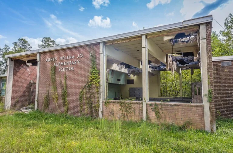 Damaged brick school building with missing parts of roof and walls, overgrown grass in front, and trees in the background.