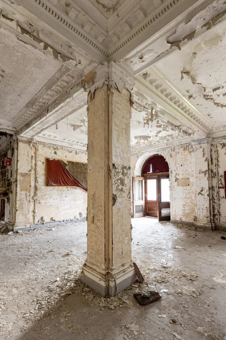 An abandoned, dilapidated room with peeling paint on walls and ceiling, a central pillar with chipped paint, debris on the floor, and a partially covered window in the background.