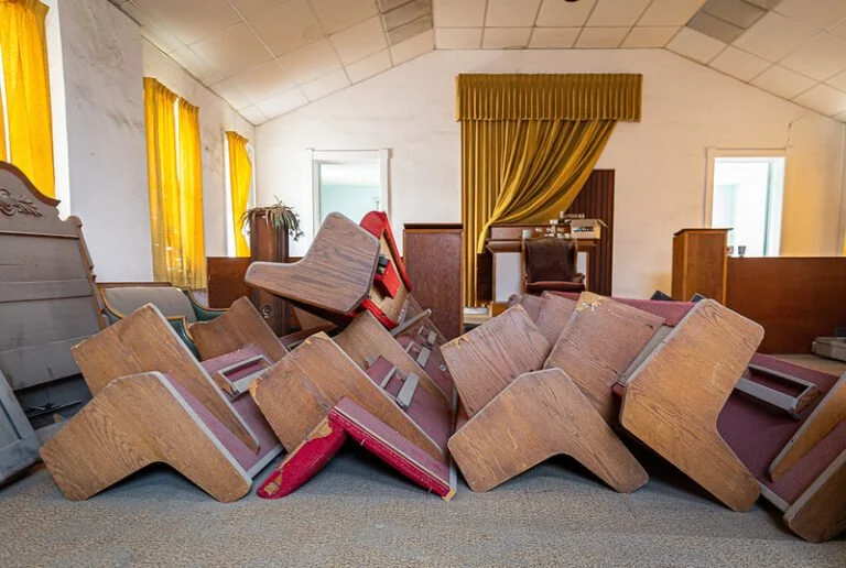 Piled-up chairs and furniture inside a room with yellow curtains and a wooden desk.