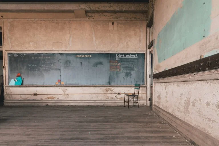 Empty classroom with a blackboard, a single chair, and some scattered objects, including a person in the corner wearing a mask.