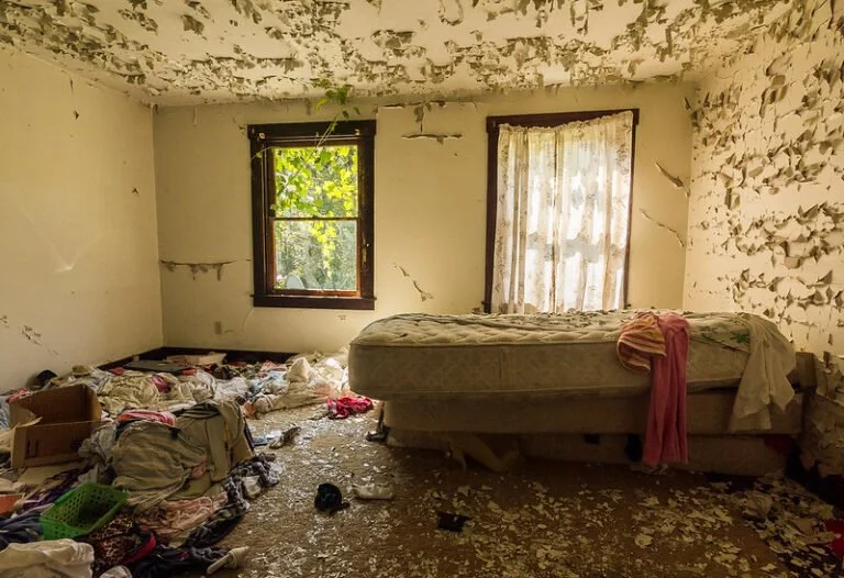 Messy bedroom with peeling paint, dirty mattress, scattered clothes, and debris on the floor, two windows with curtains letting in light.