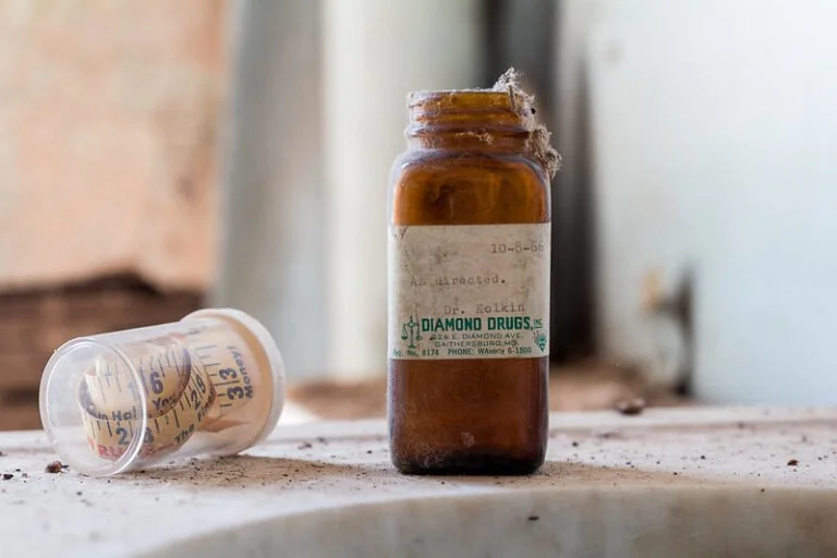 An old prescription medicine bottle and a child’s medicine cup on a dusty surface in a dusty, abandoned setting.