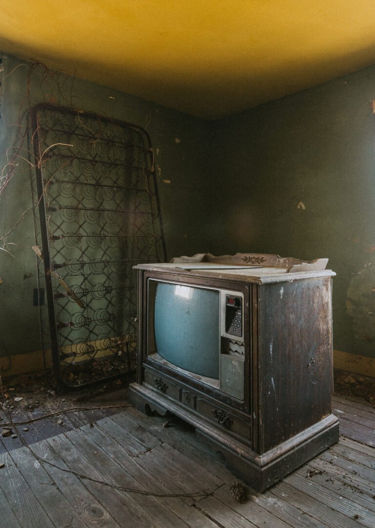 An old, dusty television on a worn wooden floor in a neglected room. Behind it, an empty metal bed frame leaning against a green wall with peeling paint, and debris scattered on the floor.