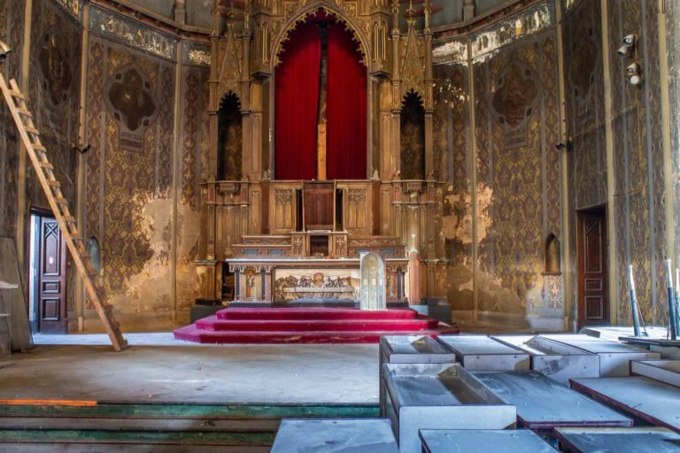 Interior of an old, abandoned church or theater with ornate gold and patterned walls, a red curtain, and a damaged stage with red carpeted steps. Debris is scattered on the floor, and some lighting fixtures are visible on the right.