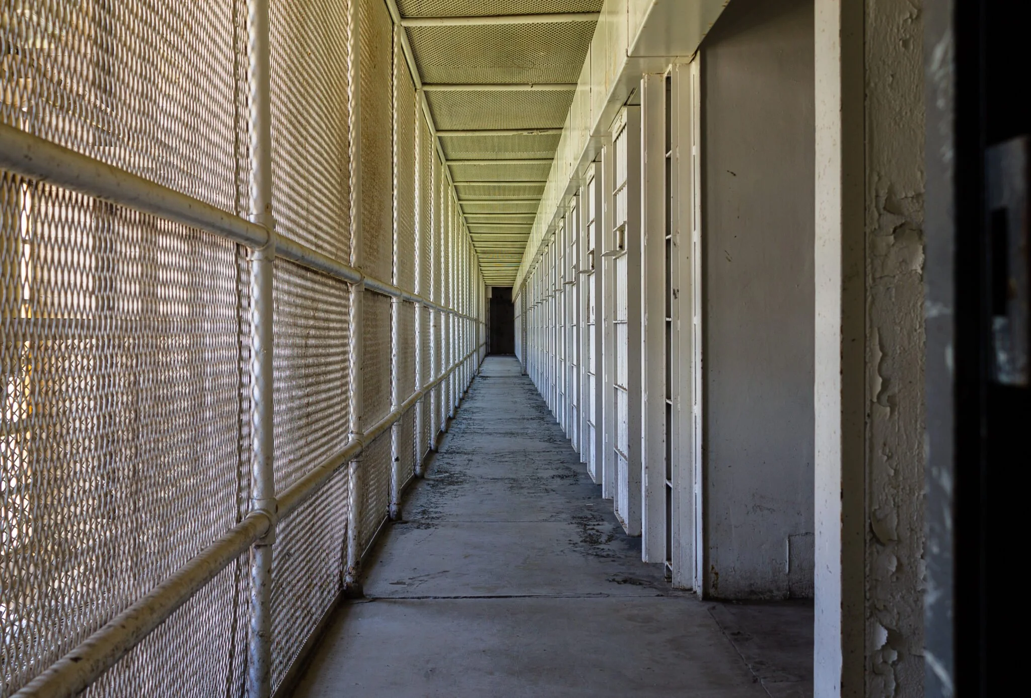 A concrete outdoor hallway in a prison or detention center with metal mesh fencing on one side and white cell doors on the other, extending into the distance.