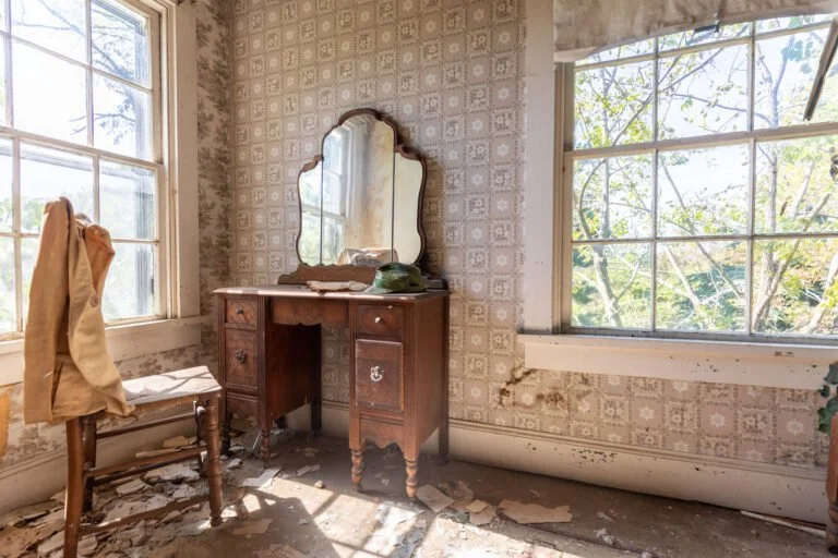 An abandoned room with peeling wallpaper, a wooden dressing table with a mirror, a vintage phone, and a wooden chair with a coat or jacket. Sunlight streams in through large open windows, revealing a view of trees outside.