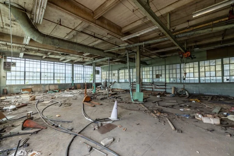 Interior of an abandoned industrial building with broken windows, scattered debris, and damaged equipment.