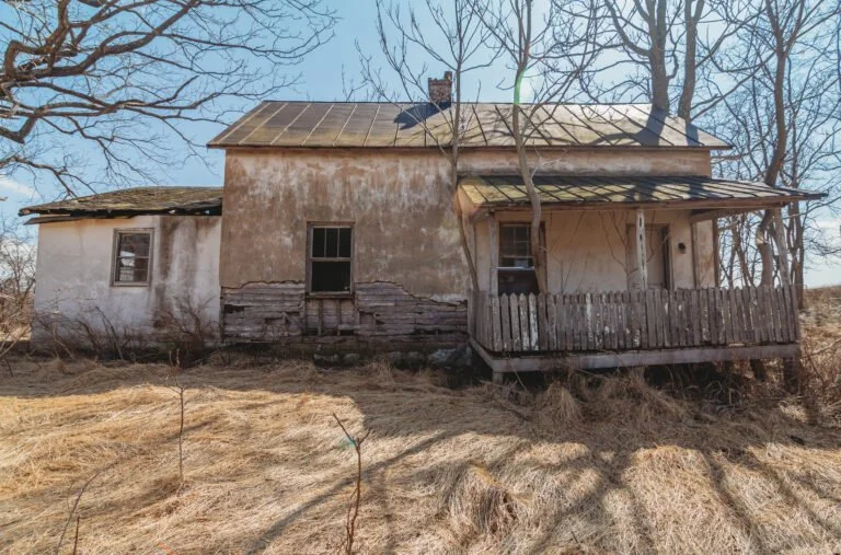 Old, abandoned house with peeling paint, broken windows, and a damaged porch, surrounded by dry grass and leafless trees.