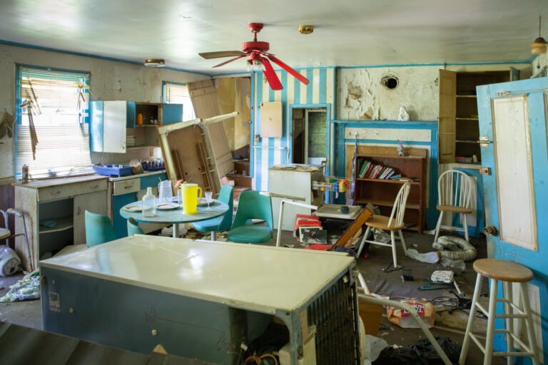 A cluttered and messy room with furniture, broken appliances, and scattered items, including chairs, a table, and various household objects.