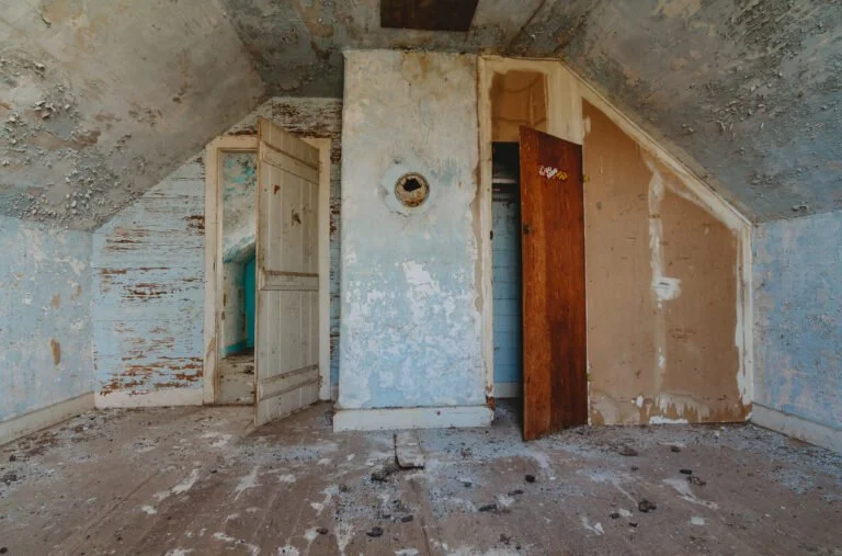 Empty, dilapidated room with peeling paint, two cluttered open doorways, and a sloped ceiling.