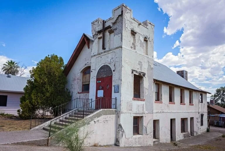 Old, abandoned church building with a cross on the front, peeling paint, and boarded-up windows, under a partly cloudy sky.