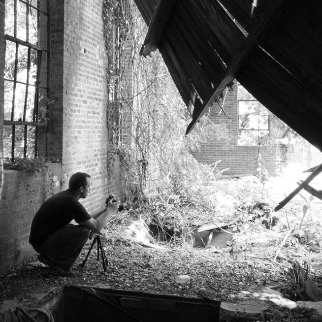 A man kneels on the ground next to a brick wall, taking a photograph with a camera mounted on a tripod. The scene appears to be in an abandoned or dilapidated building with broken windows and overgrown plants.