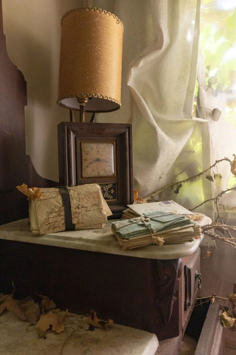 Vintage wooden clock with a floral design, a beige lamp with decorative trim, and old letters and papers tied with string on a marble-top table near a window with draped curtains, surrounded by autumn leaves.