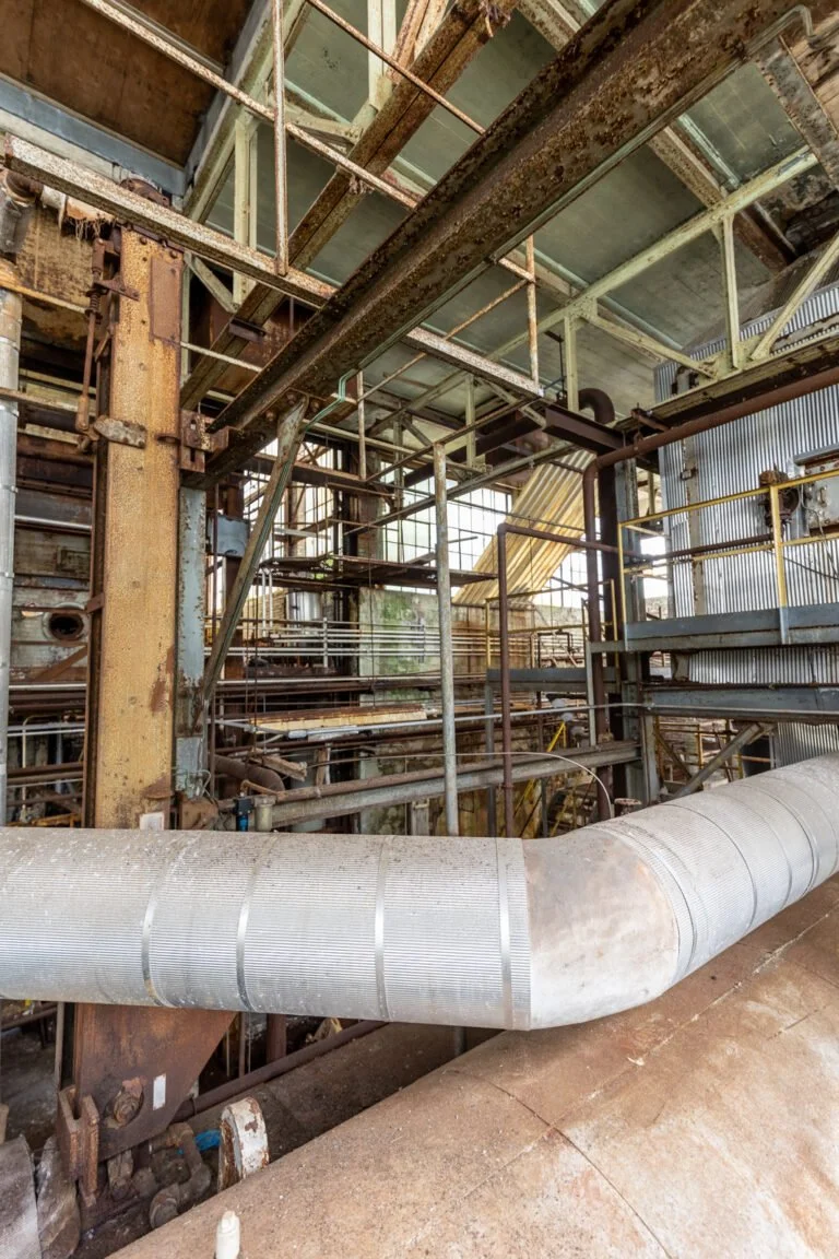 Inside an old, rusty industrial factory with corroded steel pipes, metal scaffolding, and weathered walls.