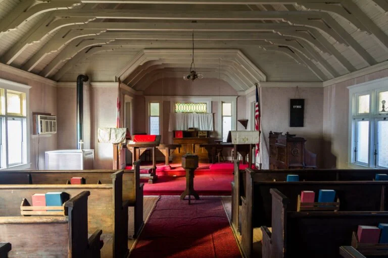 Interior of a small church with wooden pews, red carpet, and American flag. The pulpit is at the front with hymnals and a piano nearby. Windows on side walls and a small organ are visible.
