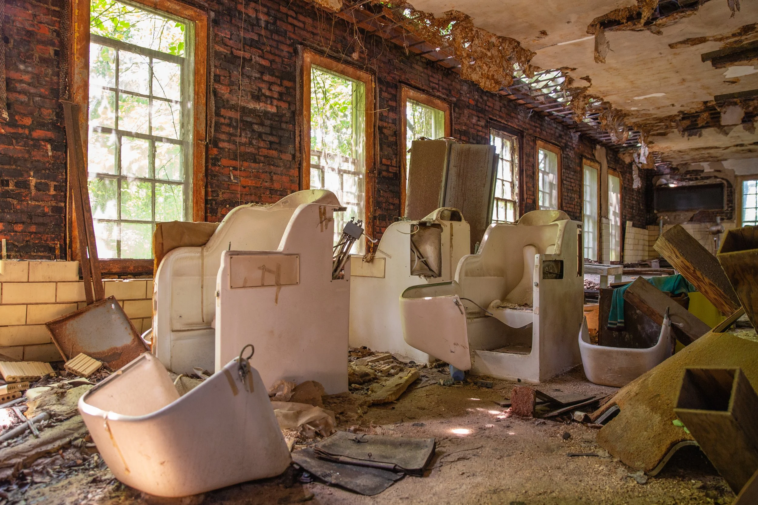 Dilapidated room with broken ceiling, peeling paint, and debris on the floor. In the foreground, there are white, broken, and abandoned commercial washing machine tubs. Multiple windows are visible, revealing overgrown outside vegetation.