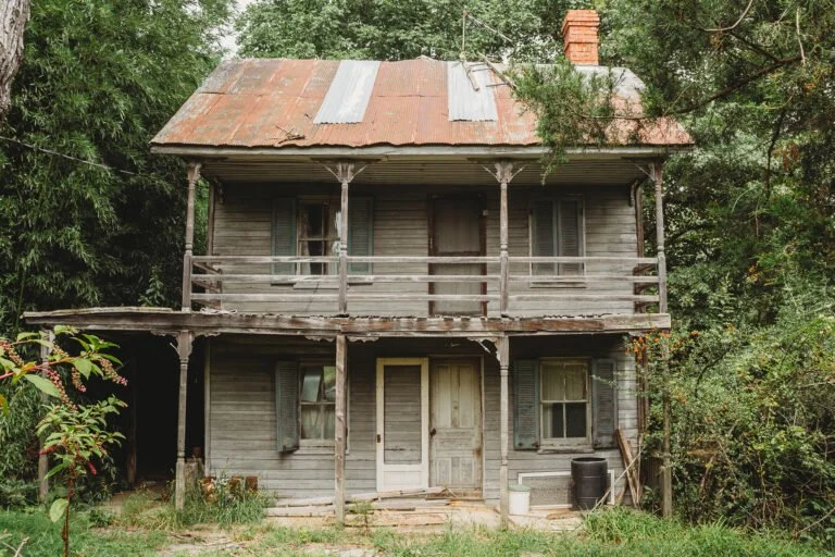 Old, weathered two-story house with a rusted metal roof, surrounded by trees and overgrown vegetation.