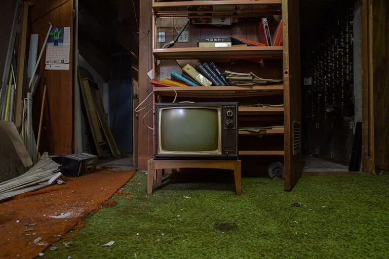 Vintage CRT television sitting on a wooden stand in a cluttered, dusty room with green carpet and shelves filled with papers and books.