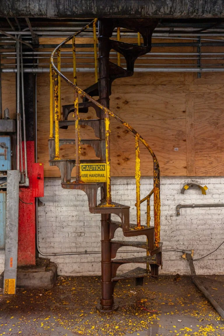An old, rusted metal spiral staircase with yellow handrails, in an industrial space, with some yellow debris on the floor.