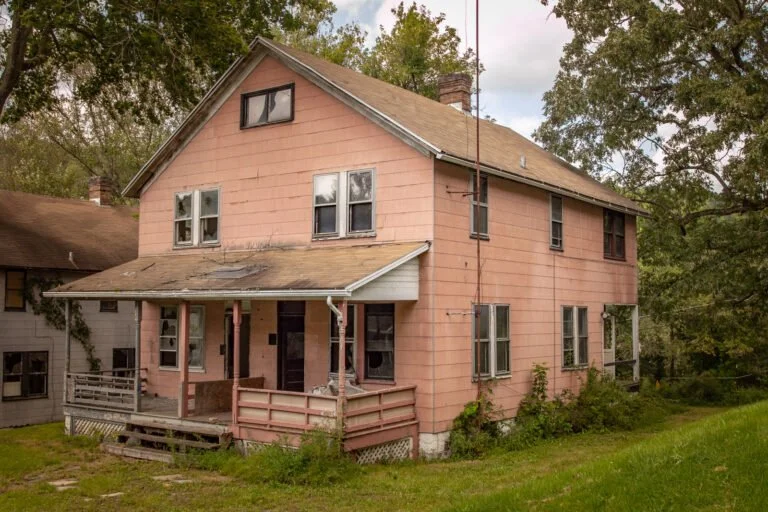 Old, two-story pink house with a front porch and broken roof, surrounded by trees and a grassy yard.