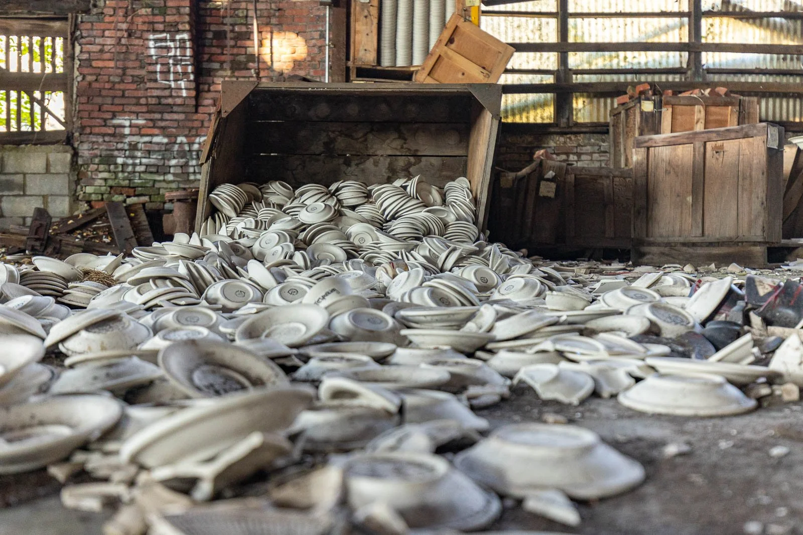 Row of broken white ceramic plates spilling out of an overturned wooden container inside an abandoned building with exposed brick walls and wooden crates.